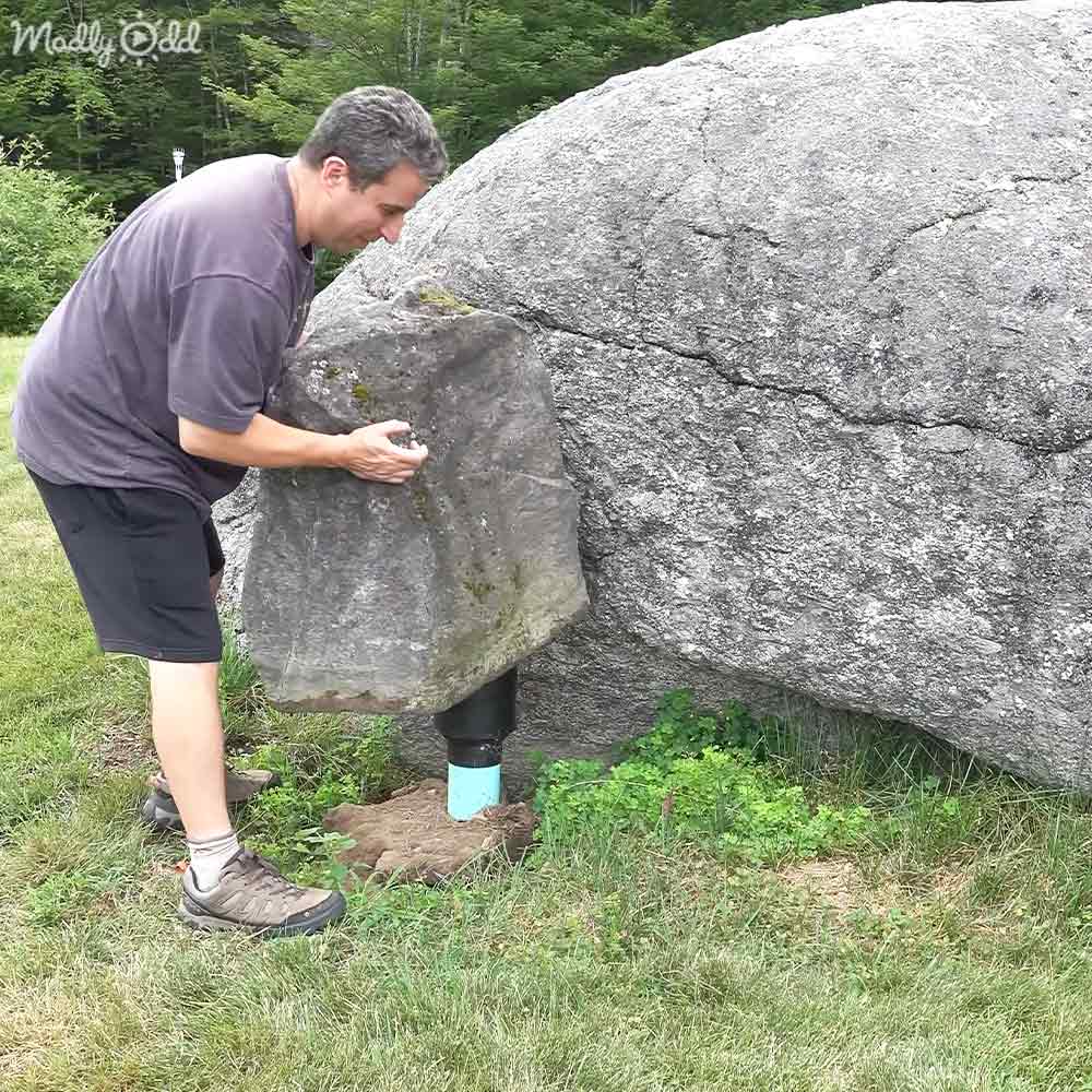 A middle-aged adult male wearing a dark purple short-sleeved polo shirt and brown hiking shoes interacts with a large split rock in a grassy outdoor setting, suggesting spring or summer. The text 'Madly Odd' is visible on the upper left corner of the image.