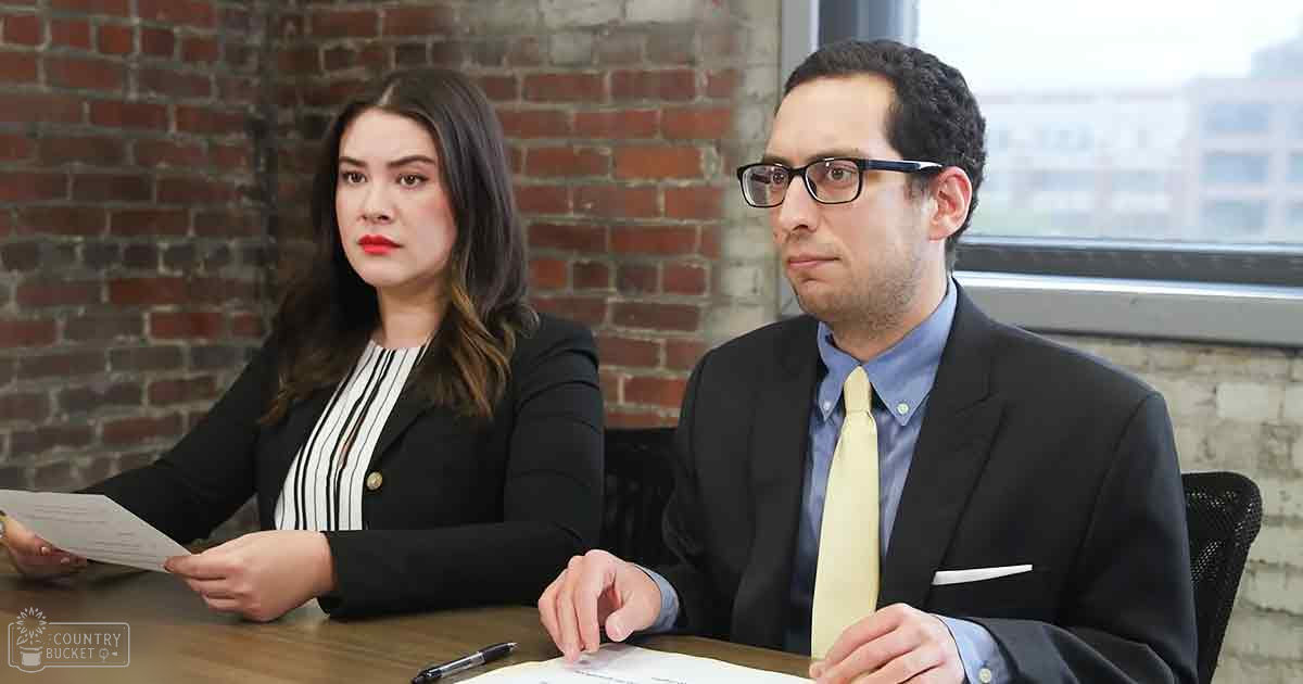 Young adults in a professional office setting with red brick walls and urban views outside, daytime lighting.