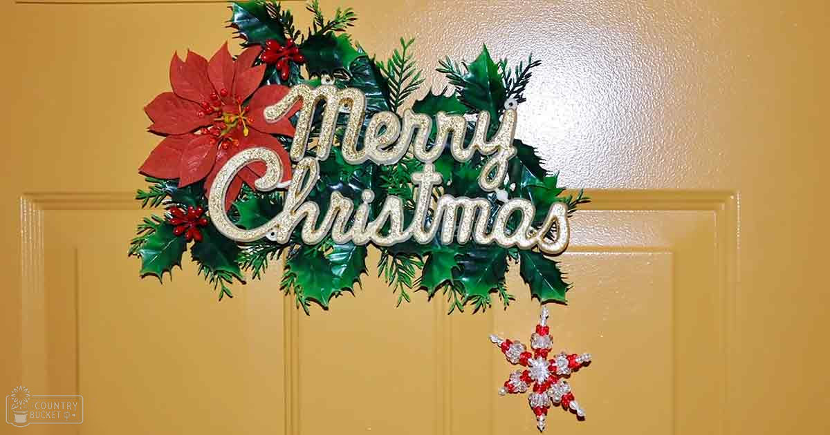 A Christmas wreath with 'Merry Christmas' written on it, adorned with green leaves, red berries, a poinsettia flower, and hanging snowflake ornament, indoors during winter.