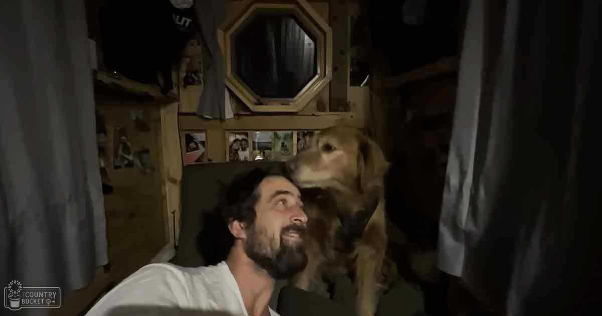 An adult male in a casual white V-neck garment indoors with a golden retriever dog, surrounded by personal belongings and wooden paneling.