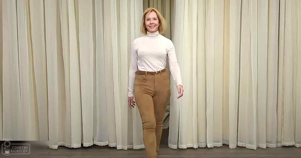 A middle-aged woman stands indoors, wearing a fitted white turtleneck sweater with brown high-waisted pants against cream-colored striped curtains.