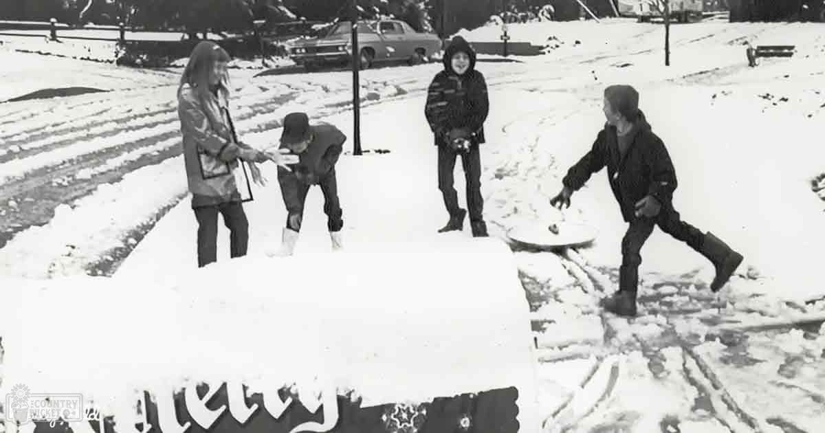 Children playing in snow-covered suburban residential area with vintage car and holiday-themed sign, winter day