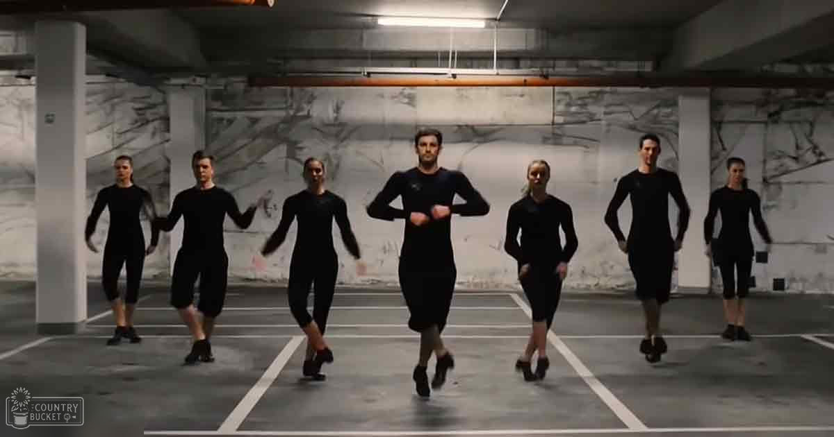 Young adult to middle-aged dancers practicing in an indoor parking garage with white pillars, a concrete floor marked for parking spaces, and walls with a marbled design under artificial lighting.