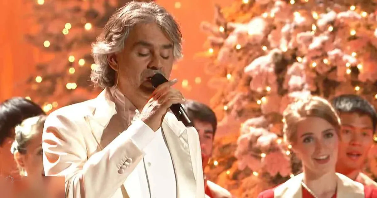 A middle-aged man sings into a microphone in an indoor performance setting with Christmas trees adorned with lights in the background, likely during the holiday season.