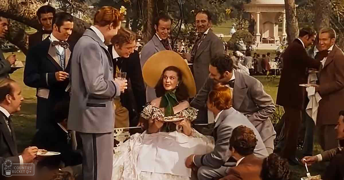 A garden party scene with an adult female in a white dress with ruffled sleeves and green scarf, surrounded by guests dressed formally in suits, under bright natural daylight amidst lush trees and a grand house in the background.