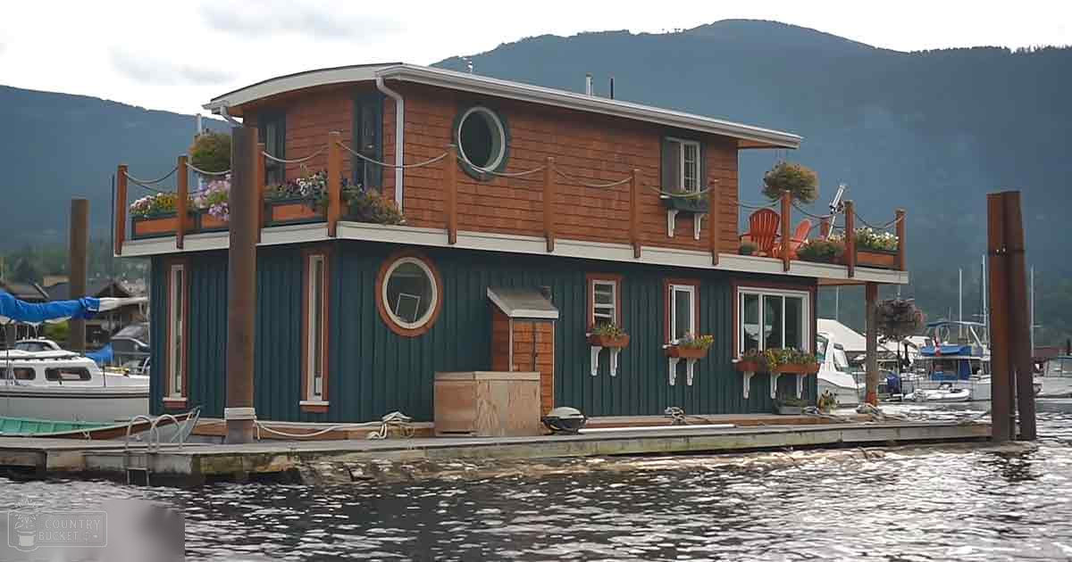 A marina scene with a two-level wooden houseboat at a dock surrounded by mountains and greenery under natural daylight.