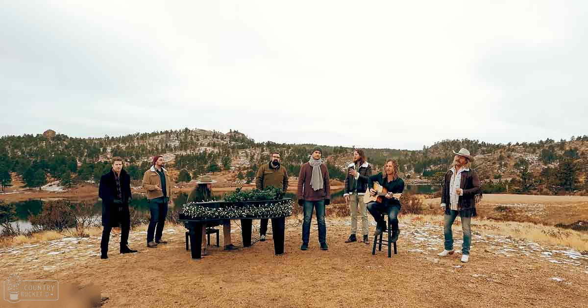 Musical group performing outdoors on a dirt path surrounded by sparse vegetation, snow patches, and a body of water; late autumn or early spring setting with earthy tones dominating the scene.