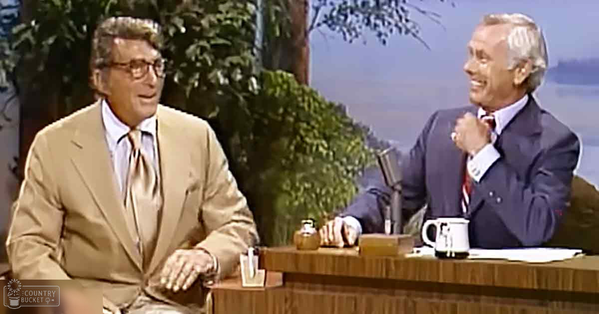 Two middle-aged adult males dressed formally, one in a beige suit jacket with a white shirt, the other in a dark blue double-breasted suit jacket and red tie, seated at a wooden desk adorned with various items including a mug and small box, against a backdrop of indoor plants suggesting a studio setting.