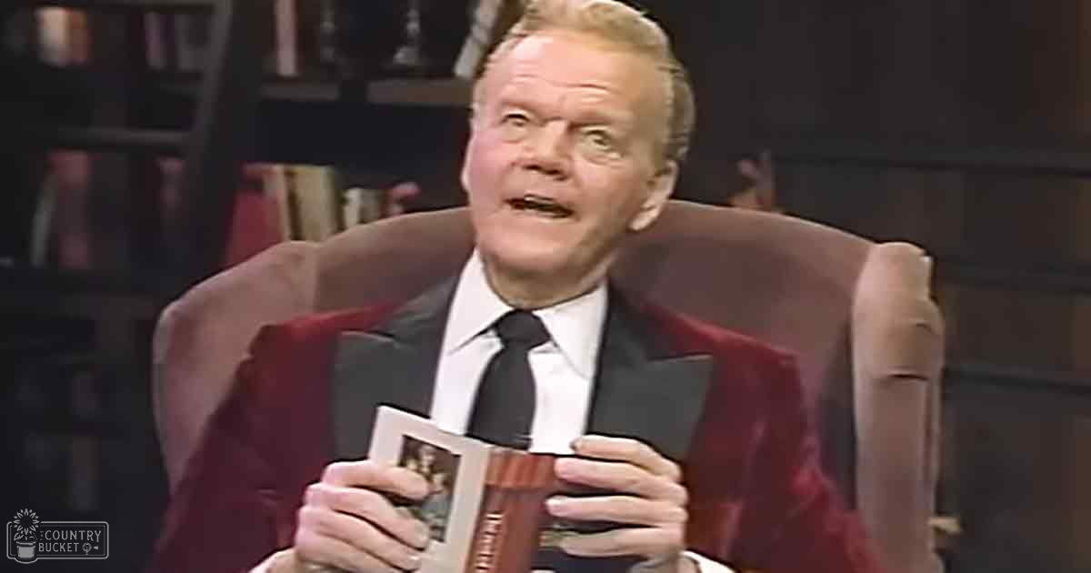 An older adult male wearing a dark suit with a red lapel, white dress shirt, and black tie sits on an upholstered armchair in what appears to be a television studio set designed for talk shows or interviews.