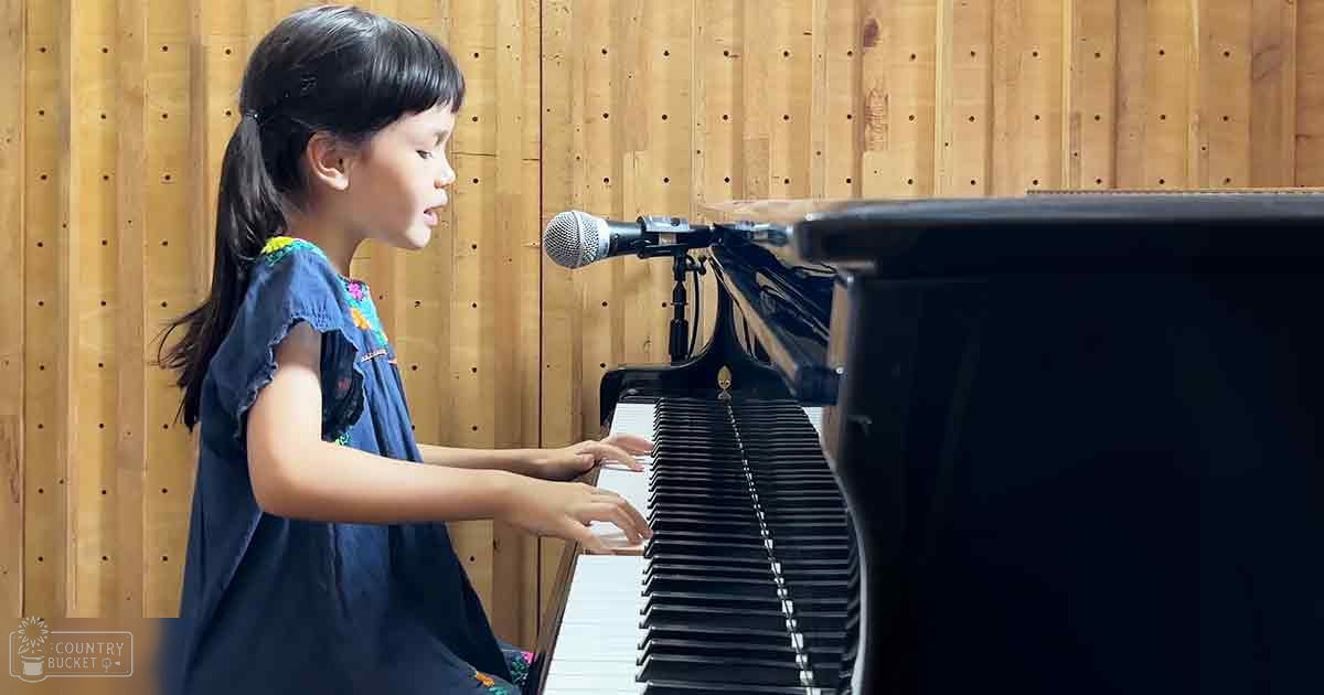 A young girl in a blue floral dress plays the piano in an indoor studio with wooden paneling.