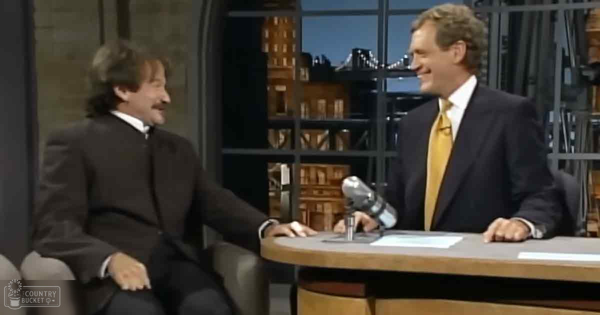 Two adult male talk show hosts wearing dark suits with contrasting white shirts and ties, seated at a round table against an urban night skyline backdrop.