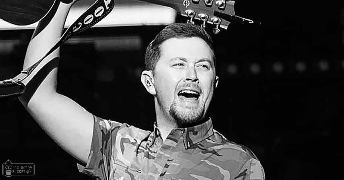 An adult male performer in a casual camouflage-patterned button-up shirt, holding a guitar above head height against an indoor concert backdrop with dim lighting and dark background.