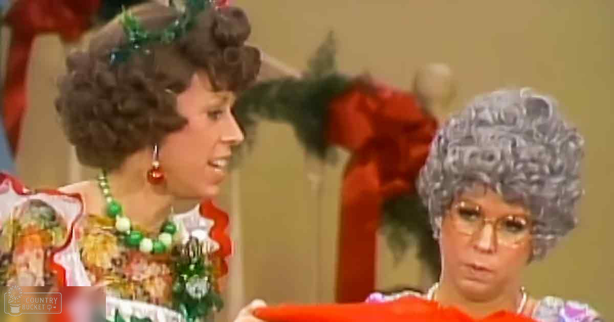 A cozy Christmas scene in a living room with two adults wearing festive attire, one in a floral red and white dress and the other in an orange patterned outfit.