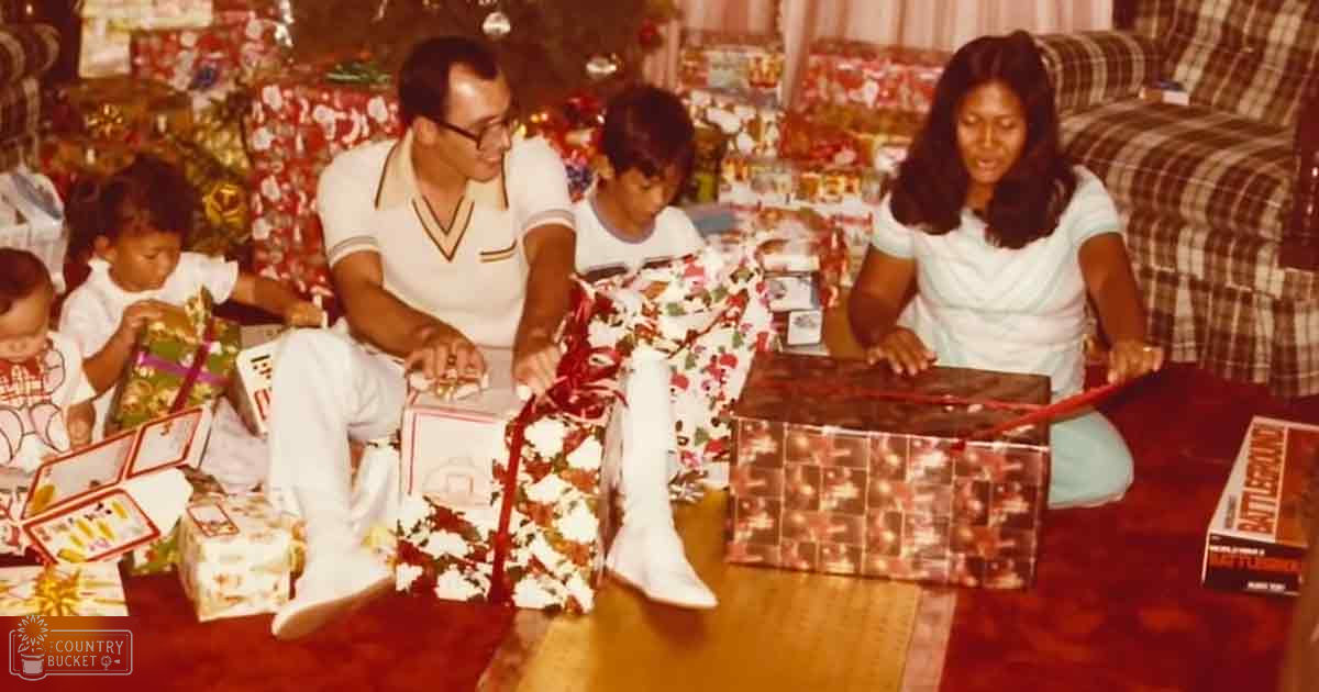 A family scene in a living room decorated for Christmas, with adults and children in light-colored holiday attire with red and green elements. The setting includes wrapped presents, a plaid-patterned couch, and artificial lighting.