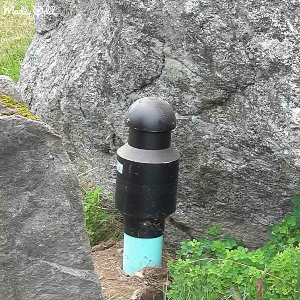 A black cylindrical object with a dome-shaped top, located outdoors near rocks in an area with light blue sections at its base and surrounded by grass and rocks.