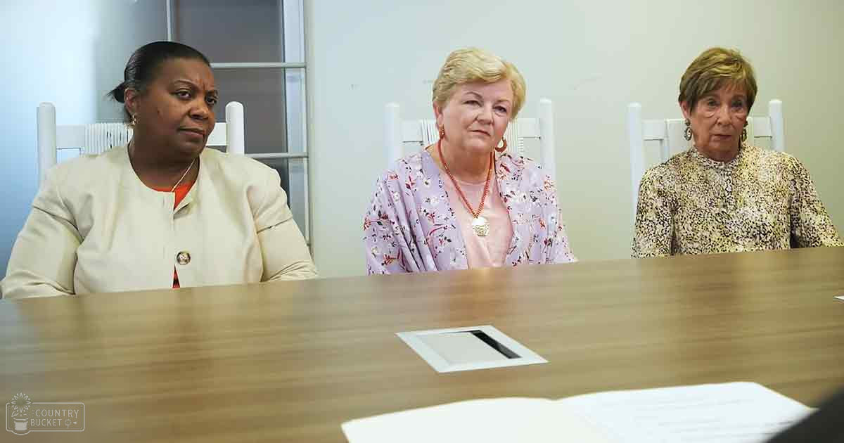 Three older adult females seated at a wooden table with vertical slat chairs, wearing light-colored blouses and patterned tops, in an office setting with natural light streaming through partially drawn blinds.