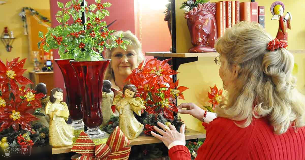 Two women in an indoor, festive setting with warm lighting; shelves filled with decorative items like books, vases, angel figurines, and poinsettia plants suggesting a Christmas or winter theme.