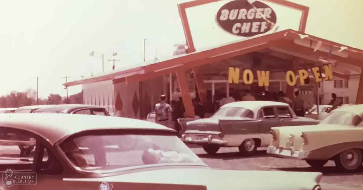 Burger Chef restaurant parking lot scene with vintage cars from the 1950s or early 1960s with muted earthy tones, trees in background and 'Now Open' sign visible.