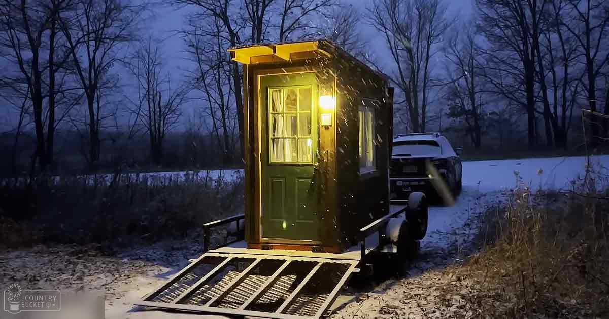 A portable cabin with a green door, yellow exterior, and interior light glowing warmly in a snowy rural landscape at dusk or dawn.
