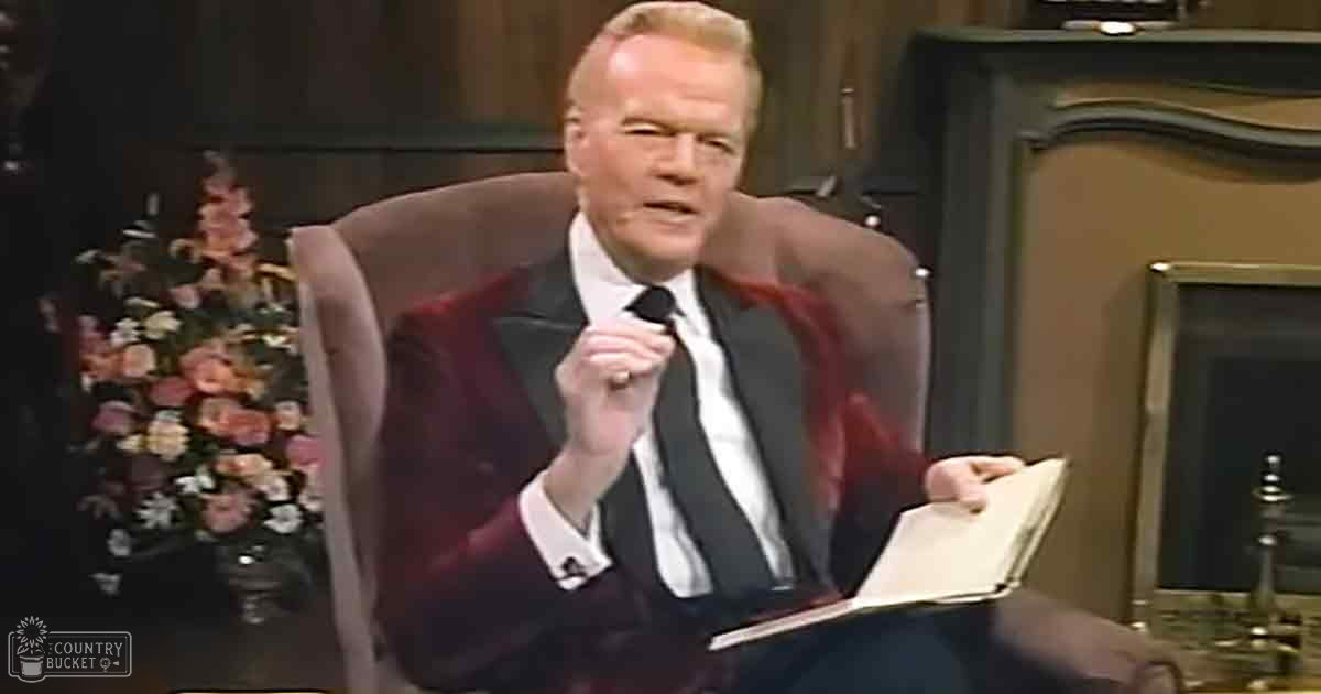 An older adult male wearing a tailored dark red blazer over a white collared shirt with a black tie, seated indoors in what appears to be a studio setup for a talk show or interview.