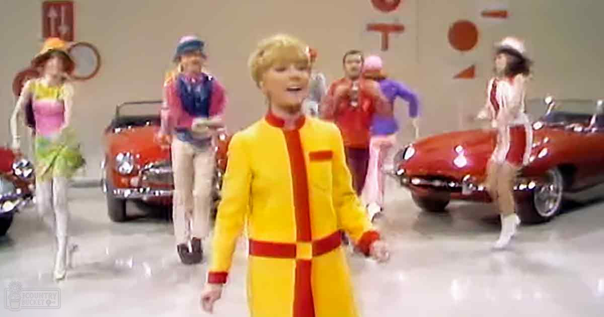 A young adult male in a yellow jumpsuit with red accents stands in an indoor setting resembling a car showroom.