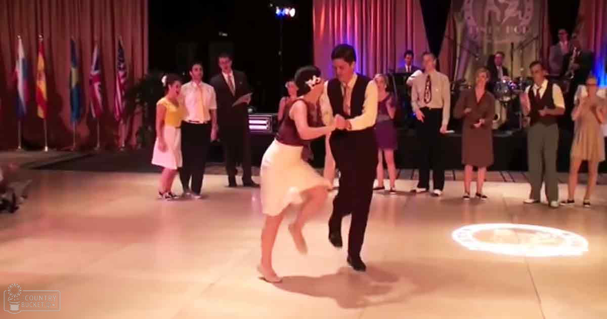 A couple dancing in a ballroom setting, with flags and musicians visible on stage. The scene is brightly lit, suggesting daytime or well-lit for an event.