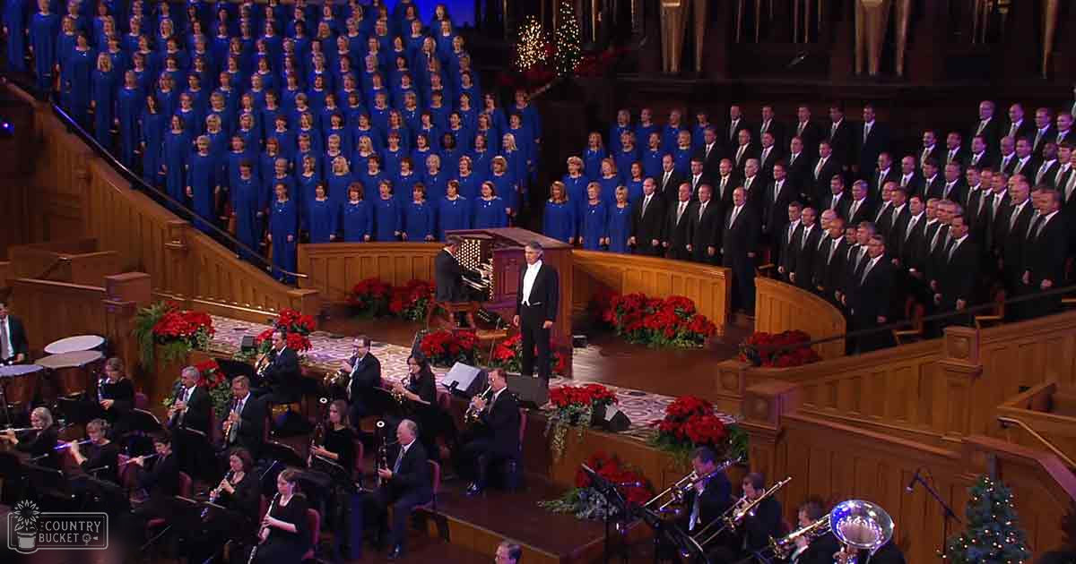 A choir and orchestra performing in a large, ornately decorated church or concert hall during the holiday season with festive red poinsettias adorning the stage.