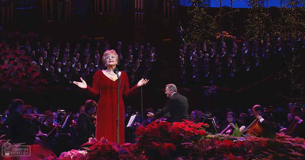 A woman in her older adult years performing on stage at a concert hall with poinsettias, suggesting it might be around Christmas time.