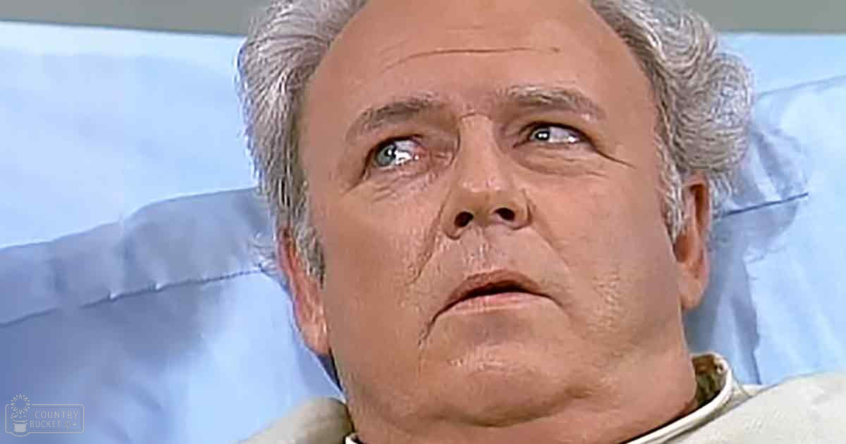 An older adult male in a hospital setting, wearing a light-colored garment suitable for medical environments. The scene is an indoor hospital room with neutral lighting and light blue sheets.