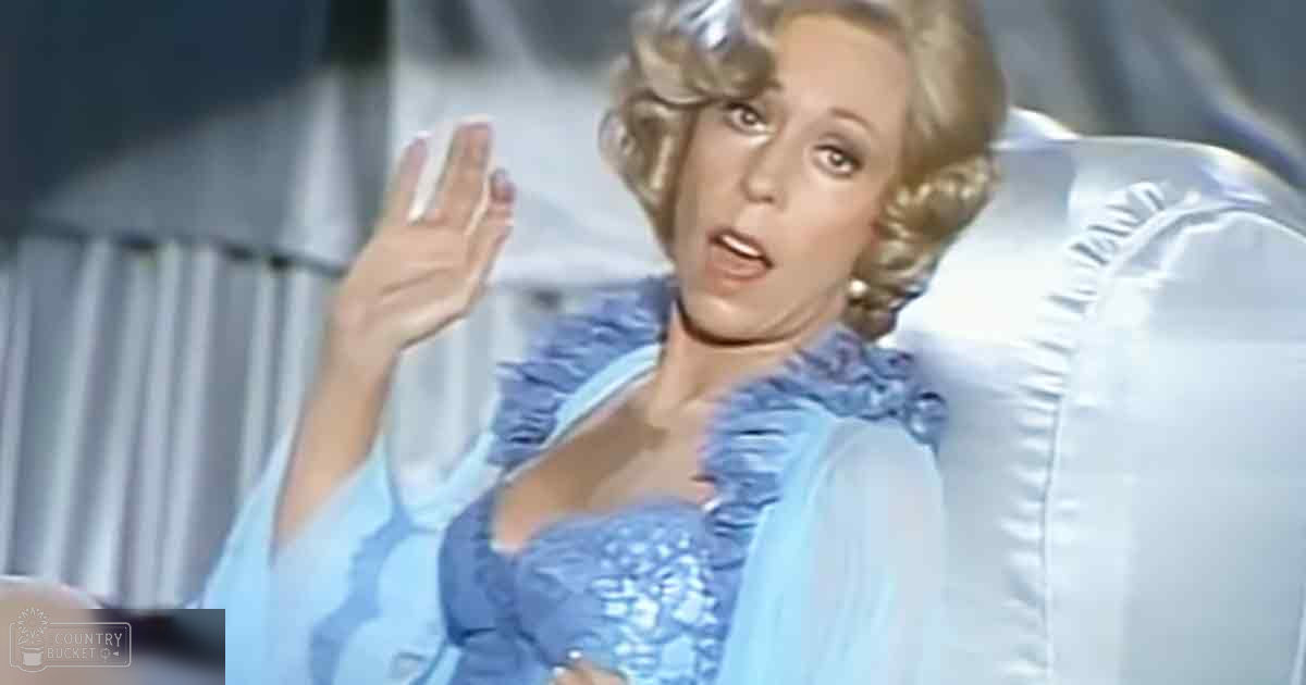 A woman in an indoor setting, wearing a mid-20th century formal dress with ruffled details and a deep V-neck.