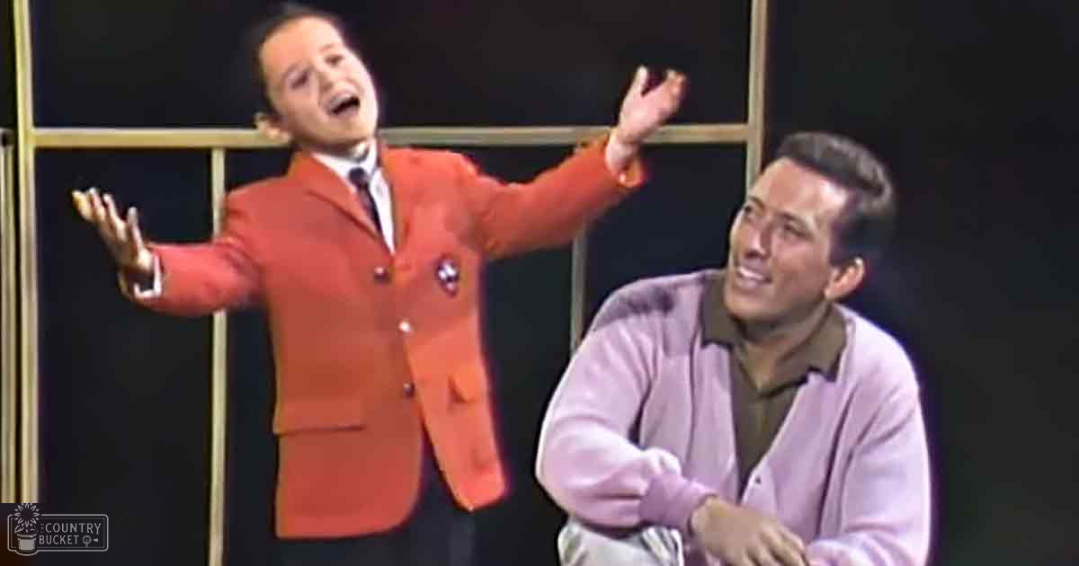 A child wearing a bright orange blazer and white shirt, standing indoors with an adult male nearby against a dark backdrop with gold lines.