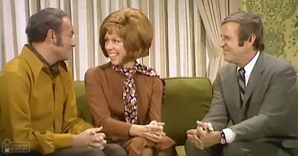 A social gathering indoors with three adults, seated on a green sofa against striped beige and cream curtains.