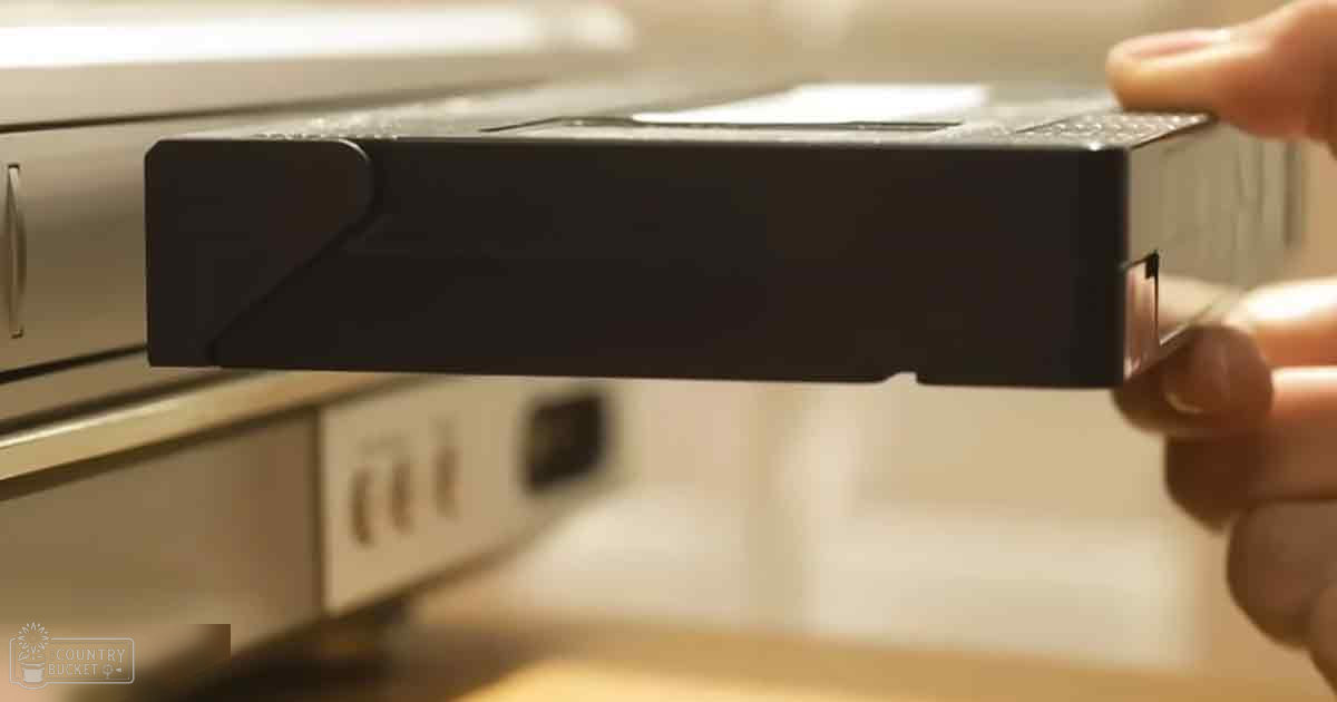 A hand holding a small black electronic device, possibly a portable charger, in an indoor kitchen setting with artificial lighting. The background is blurred but suggests cabinetry and a microwave oven.