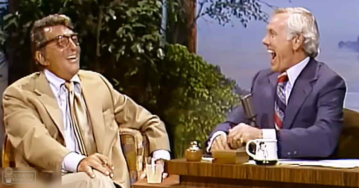 Middle-aged men in suits seated at a wooden desk with a mug and small bell on it, surrounded by an indoor studio designed to mimic an outdoor environment with visible trees and sky.