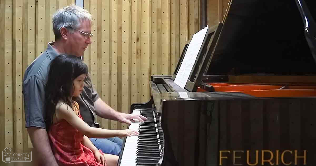 Child learning to play piano with adult instructor in indoor music studio setting, warm tones of brown and red dominate the scene.