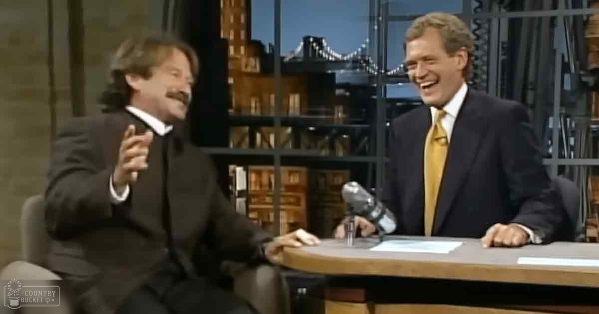 A talk show set indoors with two adult male hosts wearing dark suits against an urban backdrop suggesting New York City.