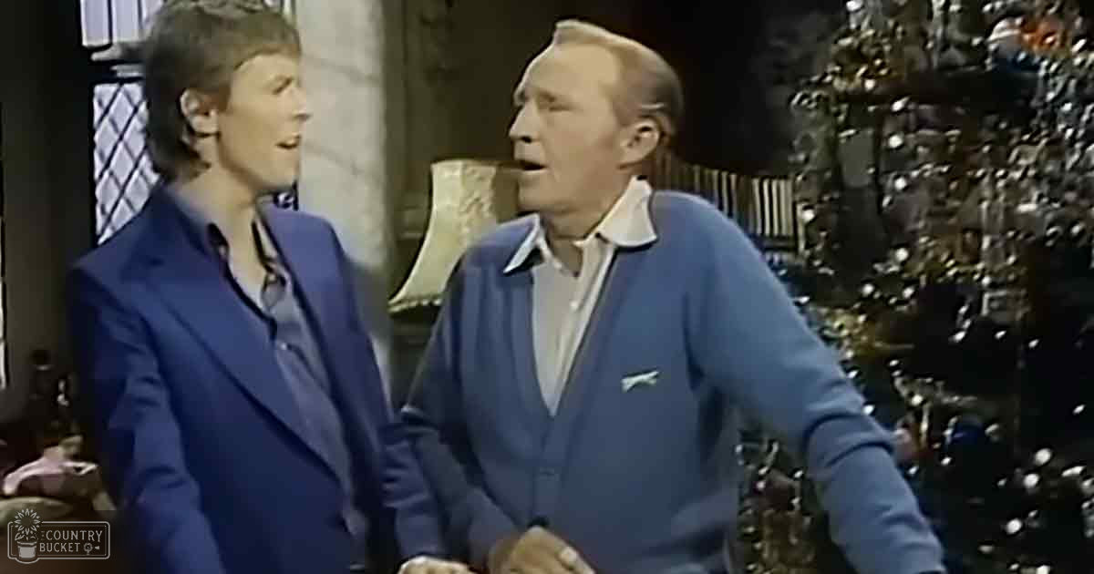 Adult female in a dark blue blazer and light-colored shirt; older adult male in a blue cardigan over a light-colored collared shirt, set against an indoor background with Christmas tree elements indicating holiday theme.