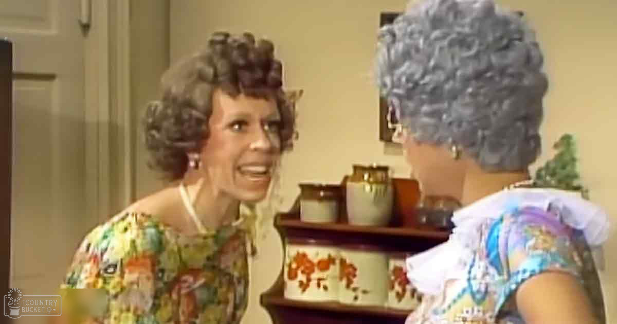 A middle-aged to older adult woman wearing a floral print blouse indoors alongside another older adult female in a white patterned blouse, with decorative items and wall decorations suggesting an indoor home environment.