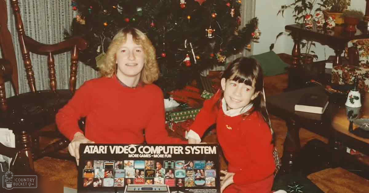 Two young girls in red sweaters in a Christmas-decorated living room with wooden chairs and an Atari box, evening light.