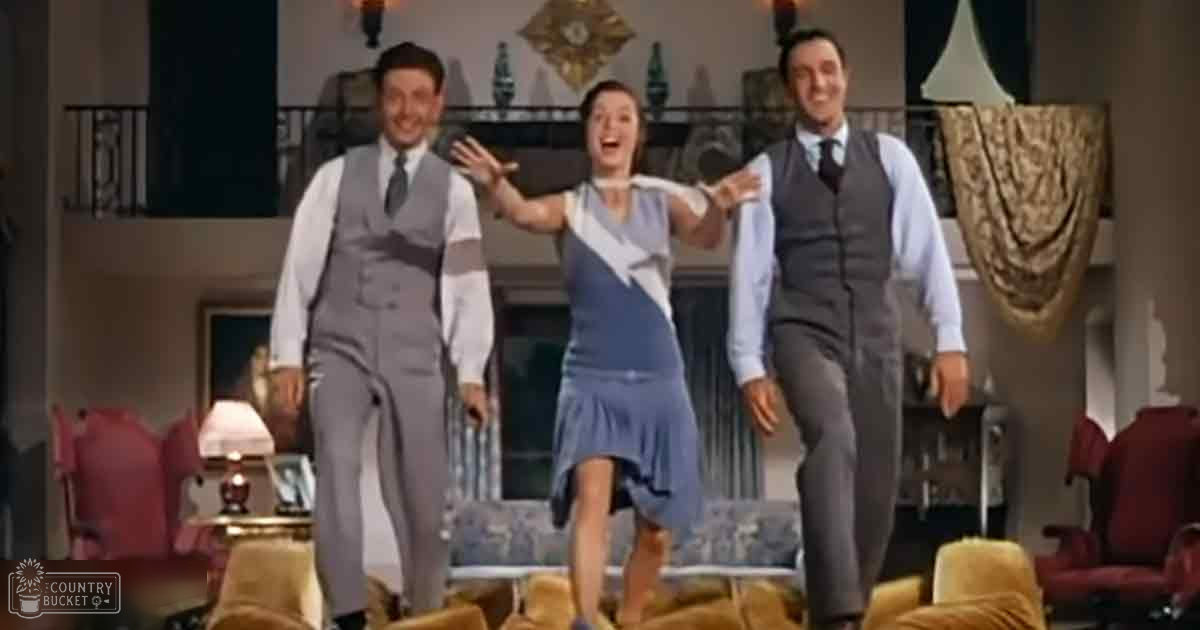 Three individuals in a classic indoor setting, wearing formal attire; adult male on left in light gray suit with white shirt and dark tie, adult female center in blue dress with ruffled hem and diagonal stripe pattern, another adult male right in similar suit design. Furniture includes red armchairs and beige sofa.