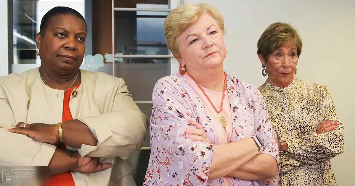Three women in professional attire, an older adult female left with a beige blazer over red top, middle-aged female center with light pink floral blouse, and older adult right in yellow and white patterned blouse, seated indoors possibly at an office or meeting room setting.