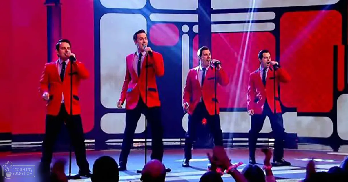 A live performance group in a stage setting with geometric shapes in red, white, and blue backdrop, adult males wearing bright red blazers and dark trousers under dramatic stage lighting.