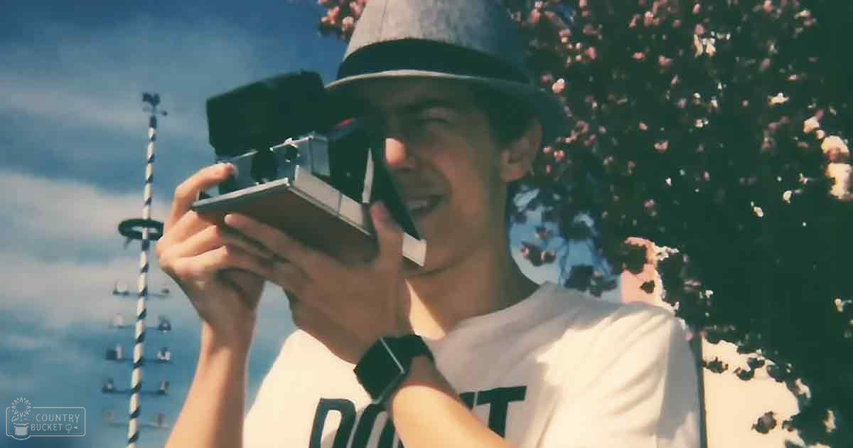 A young adult male in a relaxed white t-shirt with 'PORT' on it, holding a vintage-style camera outdoors amidst spring blossoms and partly cloudy skies.