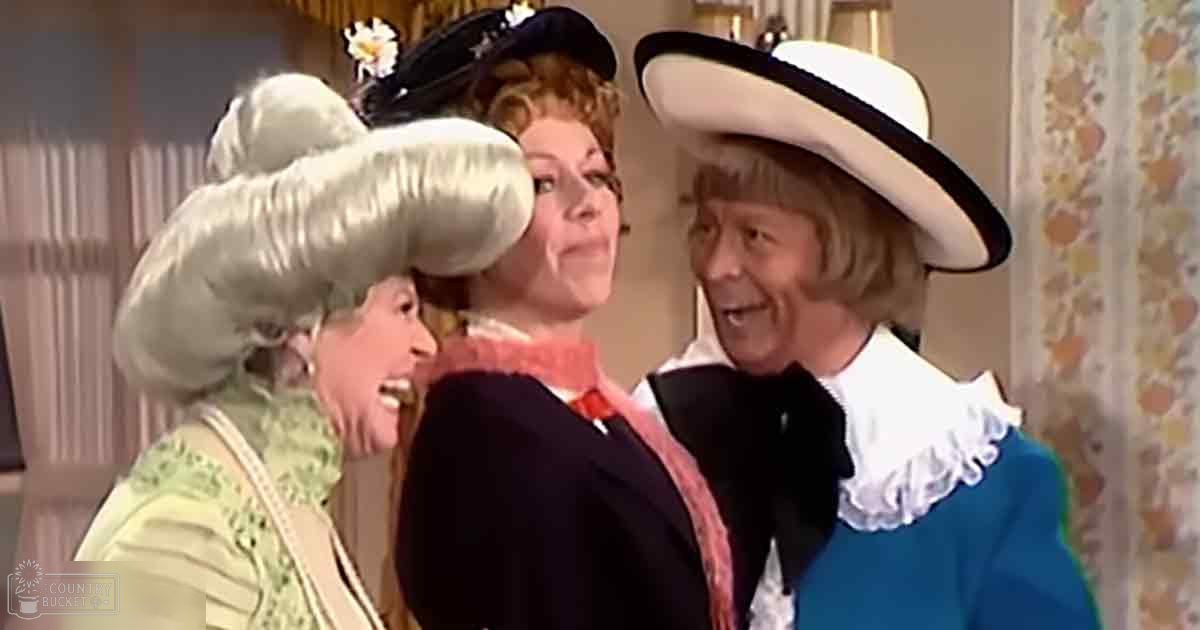 An indoor theatrical scene with three adults in elaborate costumes. The central figure wears a dark coat and floral scarf; the left, an adult female with a large white wig and green dress; the right, another older male in a blue coat and wide-brimmed hat.