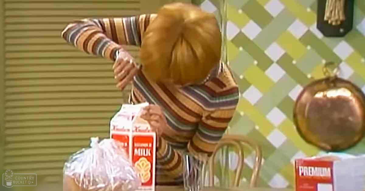 A young female child wearing a striped sweater dress stands near kitchen furniture with a checkered wall hanging in an indoor setting.