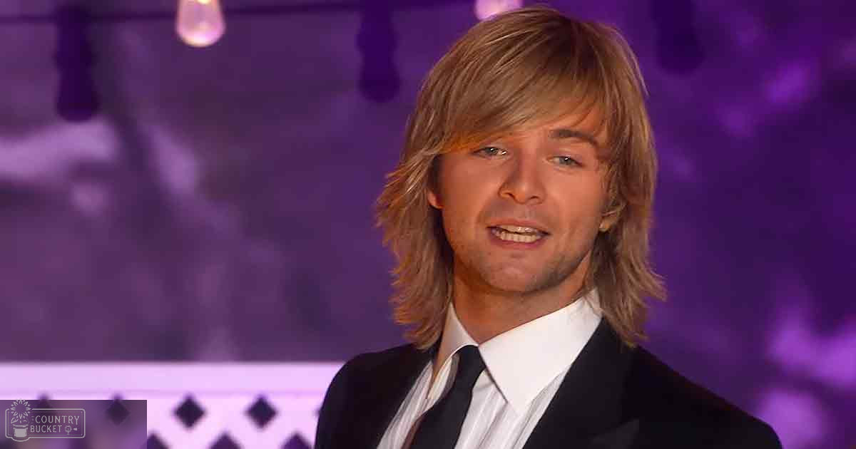 A young adult male in a formal black suit jacket with a patterned shirt, set against blurred purple stage lighting in an indoor event space.