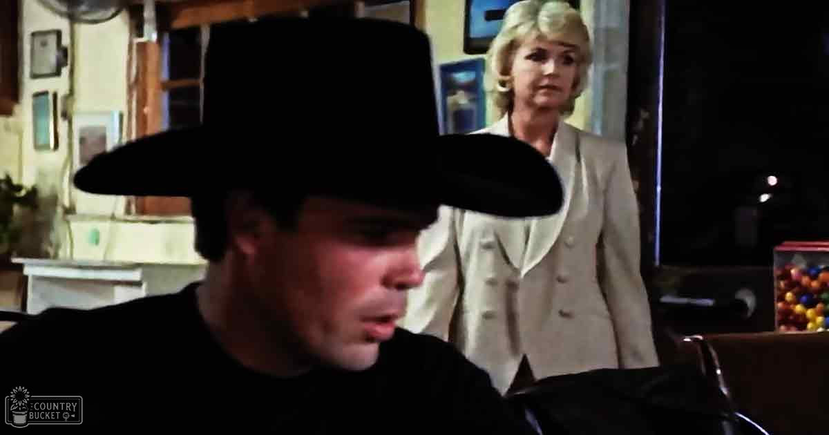 An adult male wearing a wide-brimmed black hat indoors, interacting with a middle-aged female in a cream-colored blazer. The setting appears to be a home office or study with natural daylight and various objects like a desk and colorful candies.
