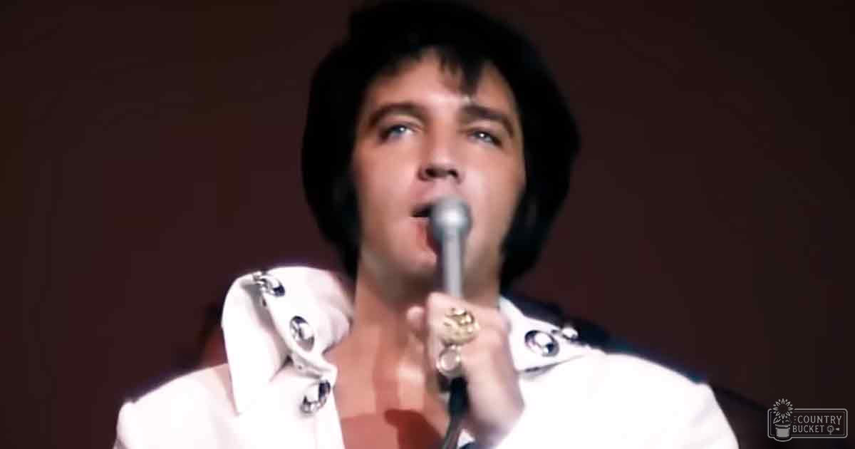 An adult male performer wearing a white jacket with silver buttons and shoulder embellishments stands under focused stage lighting at what appears to be a concert or performance venue.