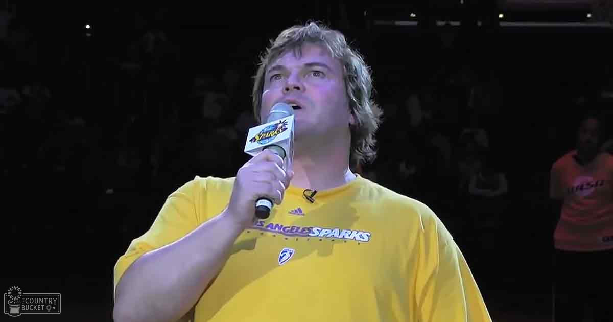 A middle-aged man in a yellow 'Los Angeles Sparks' T-shirt and holding a microphone labeled 'Spark' stands at an indoor sports arena with spectators visible in the background.