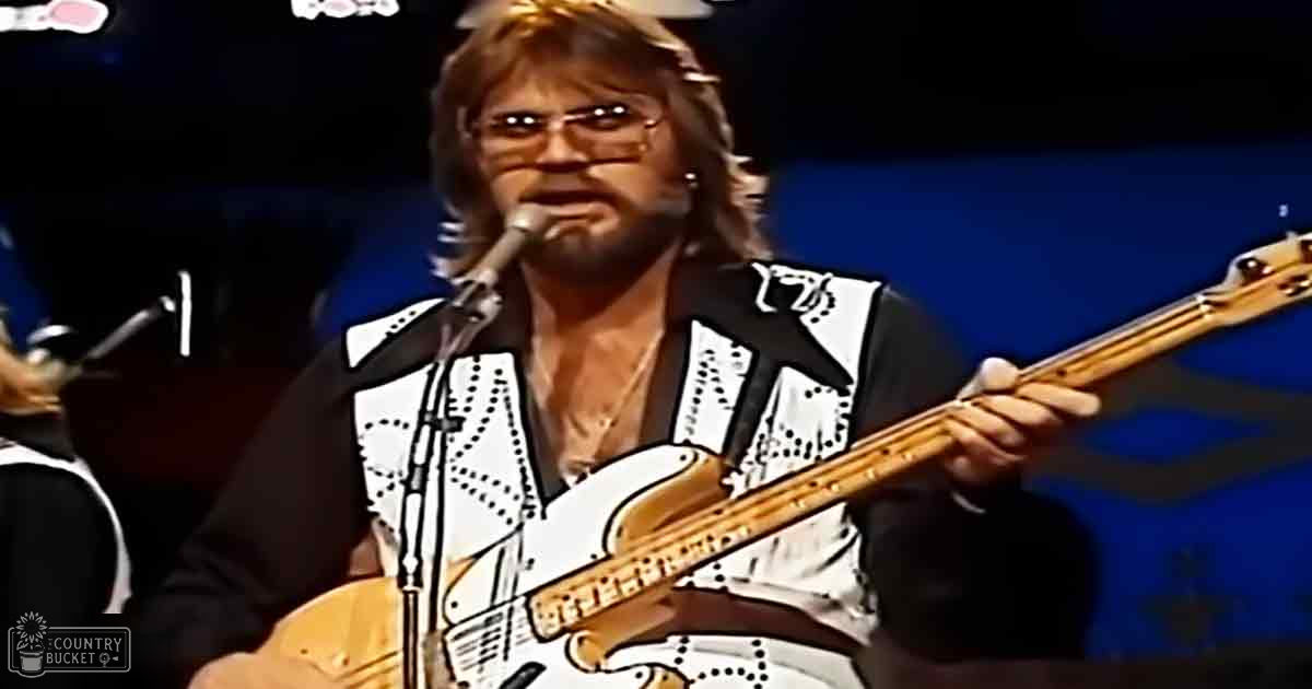 A middle-aged male performer wearing a white and black patterned vest with studded details, playing guitar in front of a microphone at an indoor concert setting.
